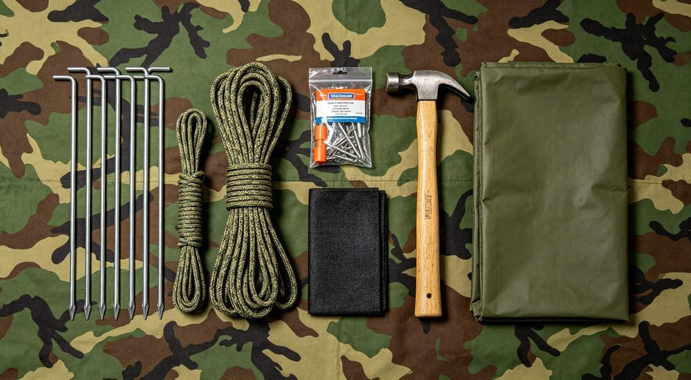 Flat lay of military tent accessories including steel stakes, guy ropes, repair kit, mallet and ground sheet organized on camouflage fabric, professional product photography