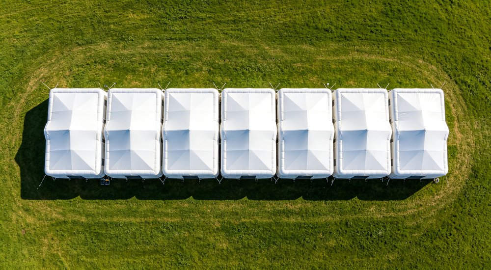 Camp expansion scene with workers adding new module to existing inflatable tent system, green grass, blue sky red and white inflatable emergency shelter for disaster relief operations Camp expansion scene with workers adding new module to existing inflatable tent system, green grass, blue sky red and white inflatable emergency shelter for disaster relief operations